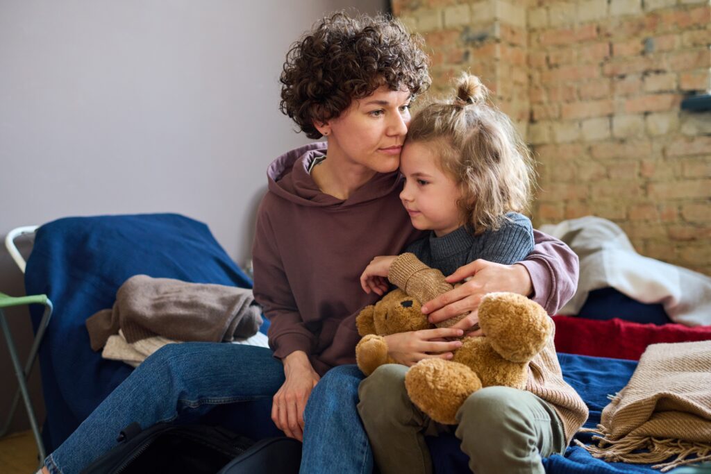 A mother sits closely with her young child on a bed in a temporary accommodation setting, both holding a teddy bear.