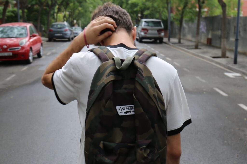 A young man walking away on a tree-lined urban street, holding his head with one hand in a gesture of contemplation or stress. He is wearing a white t-shirt and a camouflage backpack with a Vans logo. The street is quiet, with parked cars and a muted city environment in the background.