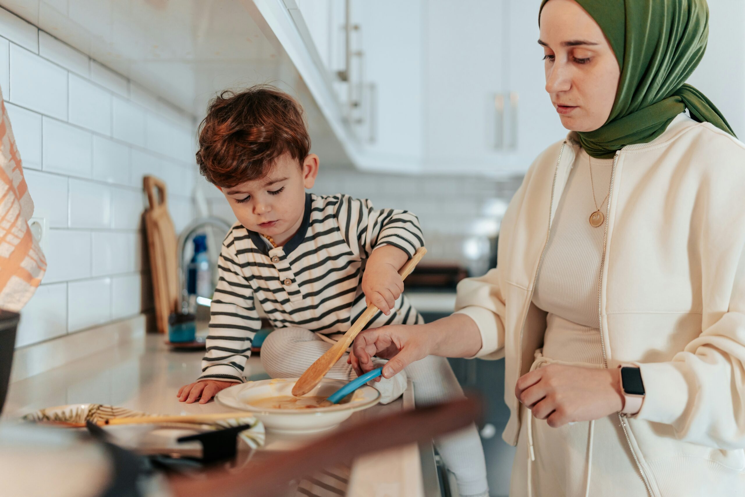 A mother wearing a green hijab and light-colored clothing is cooking in the kitchen with her young son, who is sitting on the counter and stirring a bowl with a wooden spoon. The kitchen has a white tile backsplash and white cabinets.